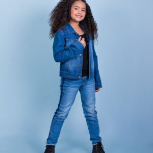 Smiling girl in denim jacket and jeans standing confidently against a blue backdrop.