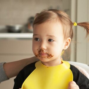 Cute toddler with chocolate-covered face wearing yellow bib in a cozy kitchen.