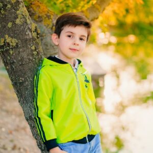 A young boy stands by a tree, wearing a vibrant jacket, with an autumn backdrop.
