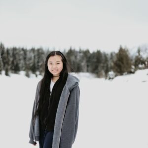 A smiling Asian woman in a gray jacket standing amidst a snowy winter landscape.